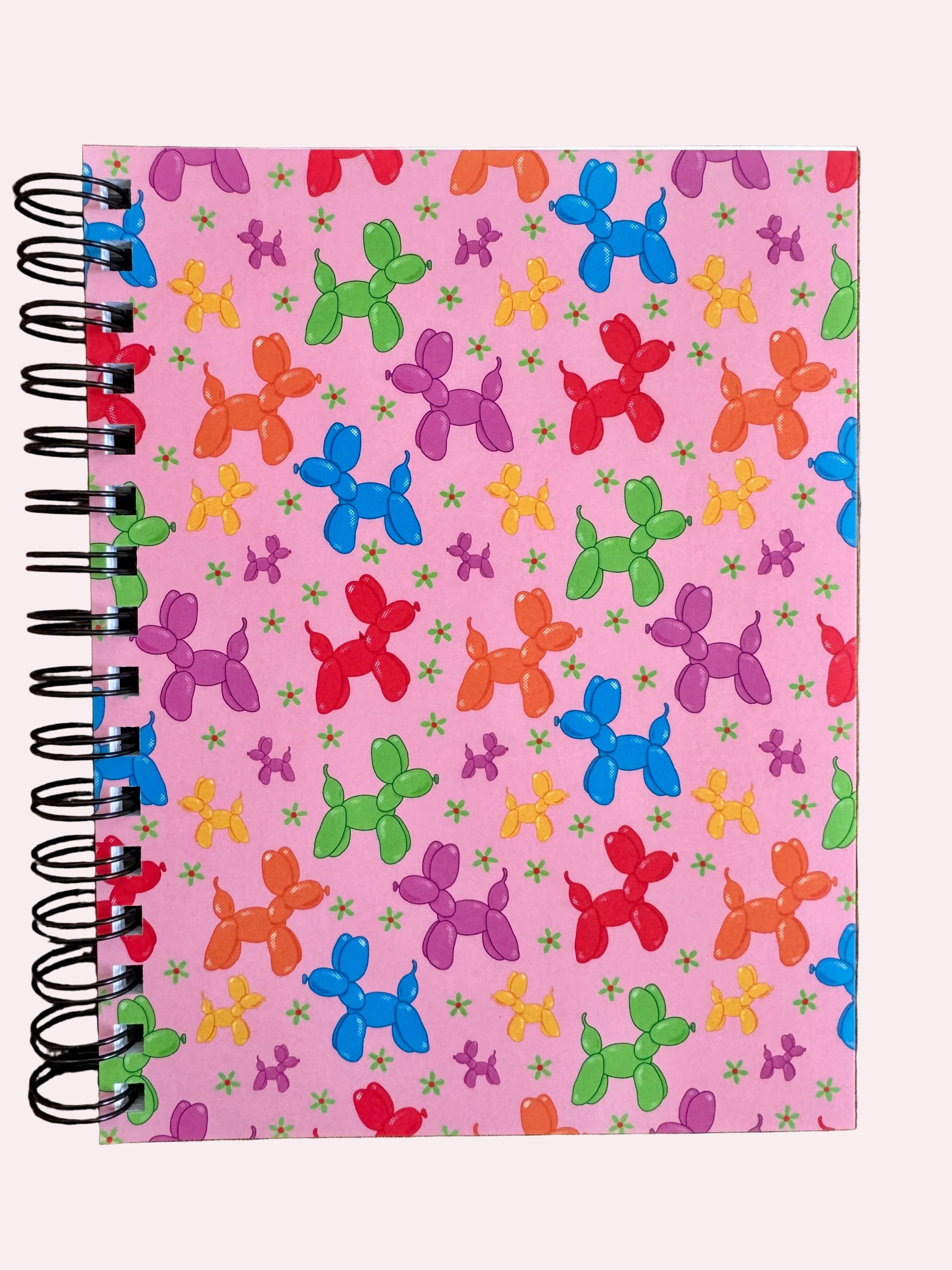Notebook with colorful balloon dog pattern on a pink background, placed on a wooden surface.