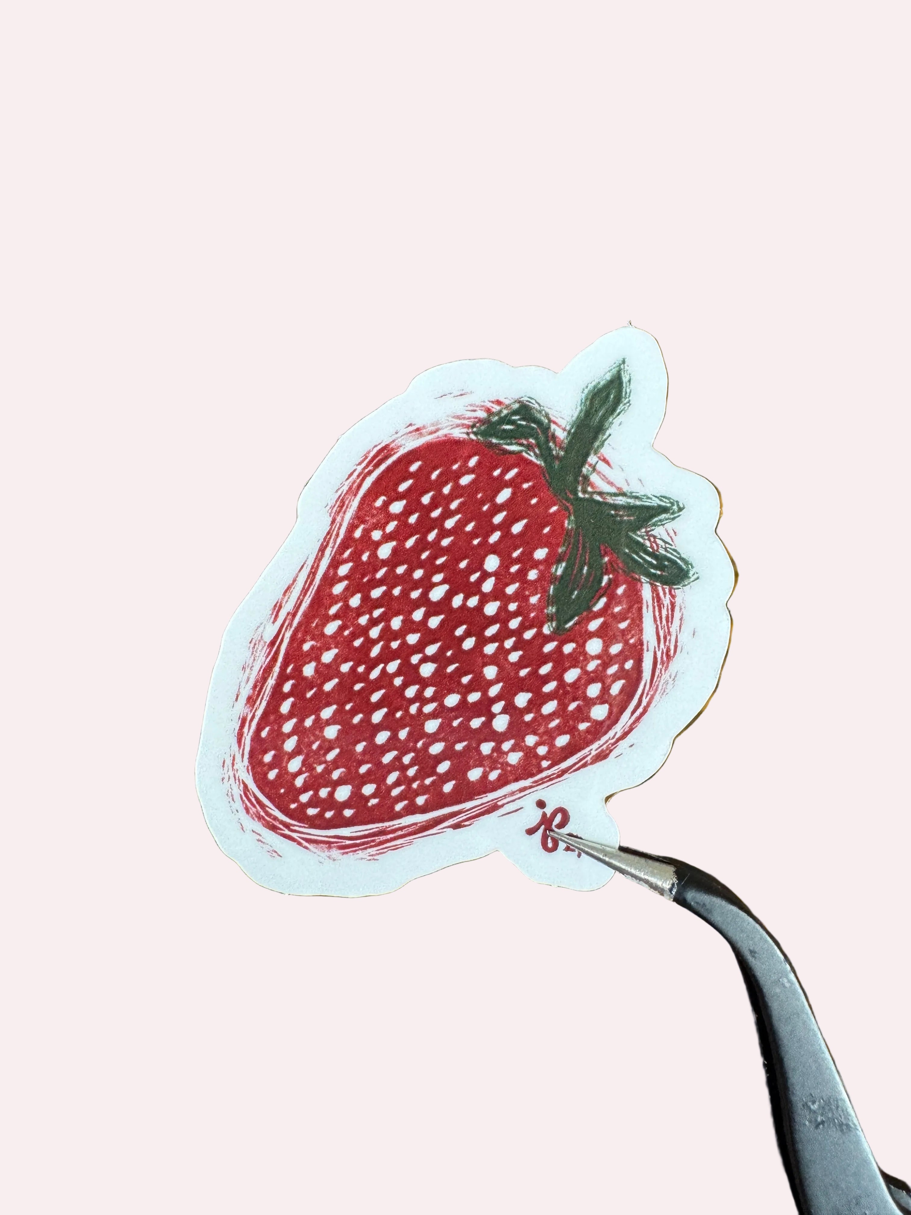 A die-cut sticker of a red strawberry with green leaves, held by tweezers over a wooden surface.