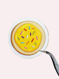 A round sugar cookie decorated with bright yellow icing and colorful sprinkles, placed on a white plate over a dark wood grain background, with a pair of tweezers holding the edge of the plate.