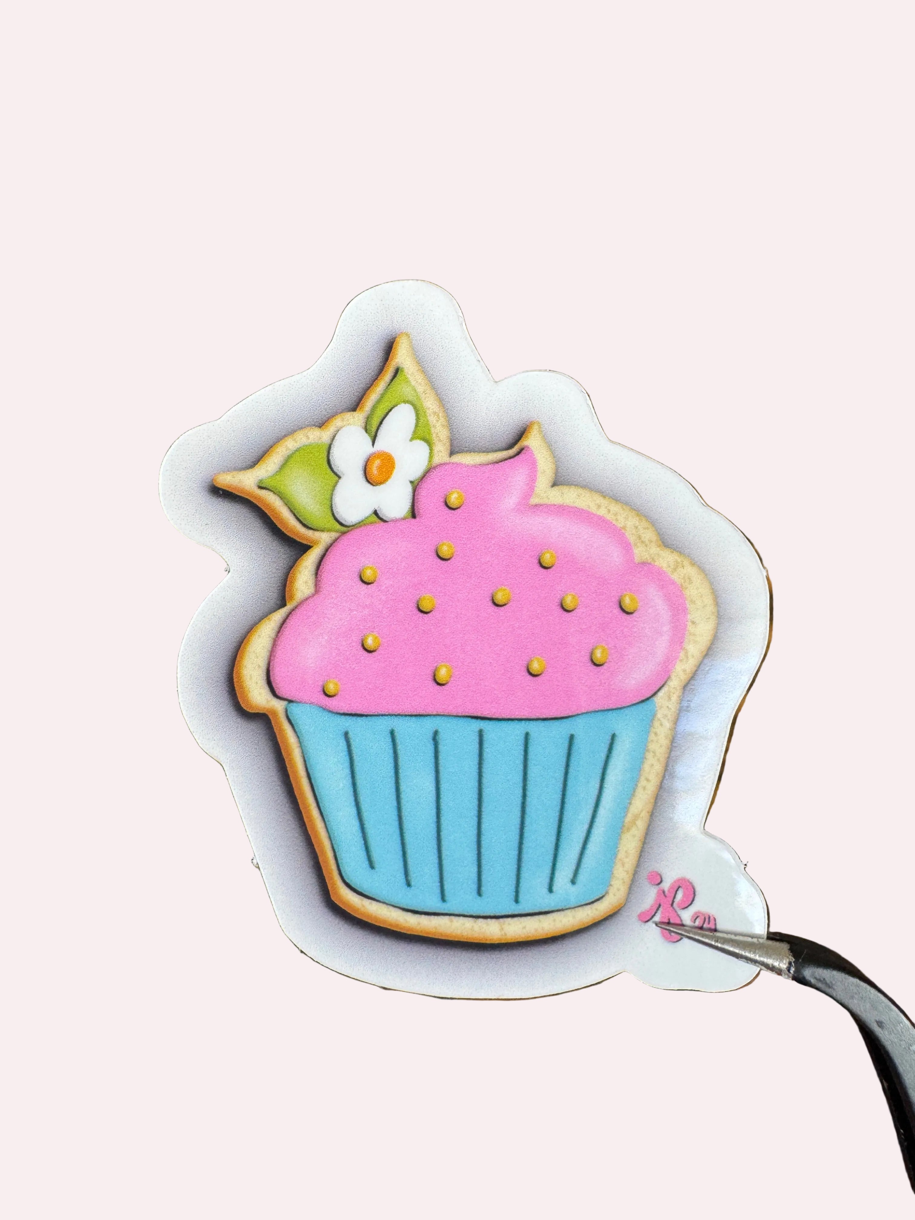 A close-up of a decorated cupcake-shaped cookie or sticker with pink frosting, blue frosting, gold sprinkles, green leaves, and a small white flower, shown on a wooden surface with tweezers holding the top edge.