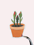A die-cut sticker depicting three green cactus-like plants in a terracotta pot, held by tweezers over a dark wood grain background.
