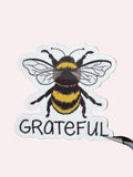 A die-cut sticker featuring a detailed illustration of a bumblebee next to the word 'GRATEFUL' in black lettering, displayed on a brown wood-grain surface.