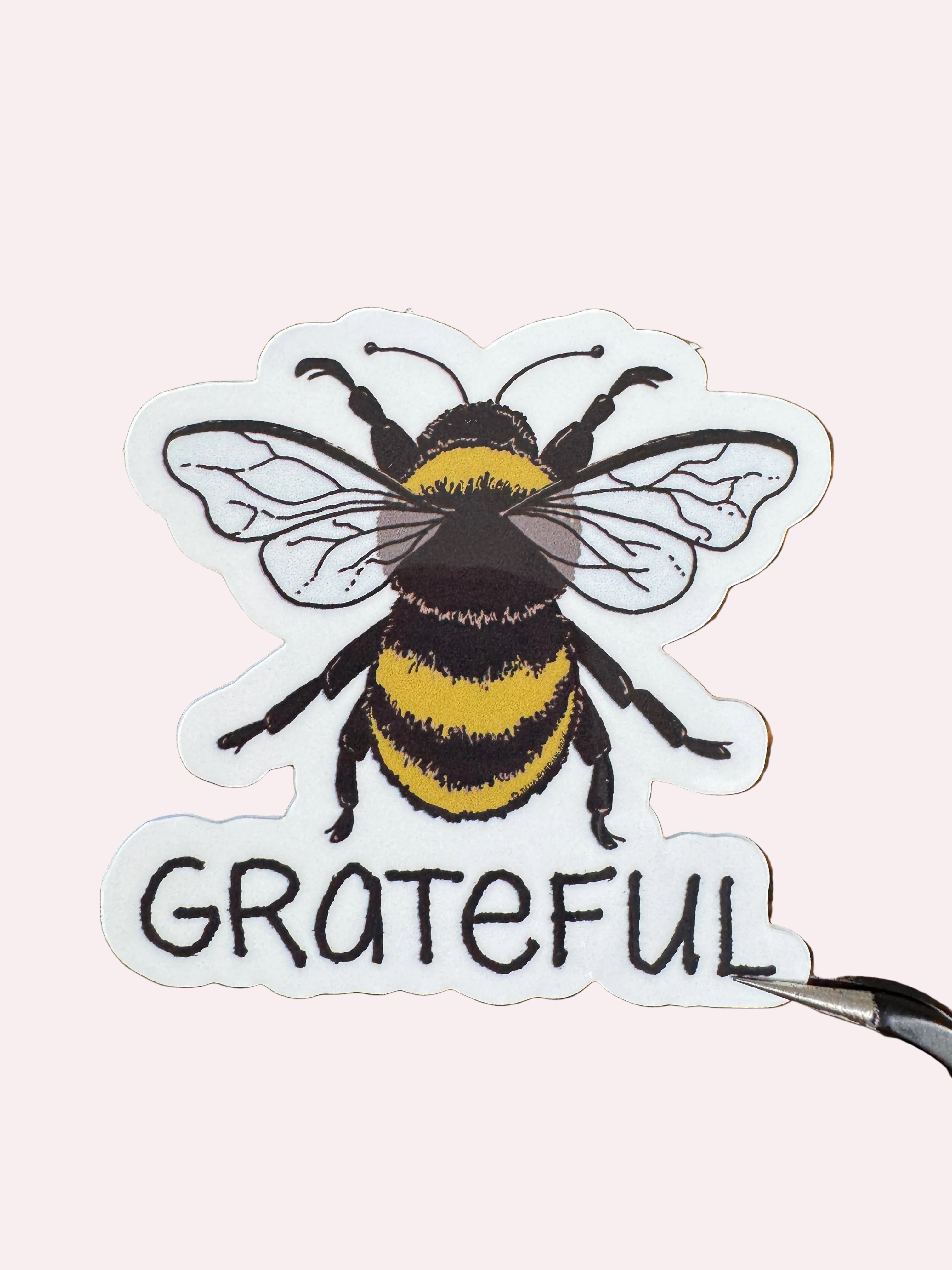 A die-cut sticker featuring a detailed illustration of a bumblebee next to the word 'GRATEFUL' in black lettering, displayed on a brown wood-grain surface.