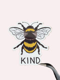 A die-cut sticker featuring a detailed bumblebee and the word 'KIND' in black lettering, held by tweezers on a dark wood background.