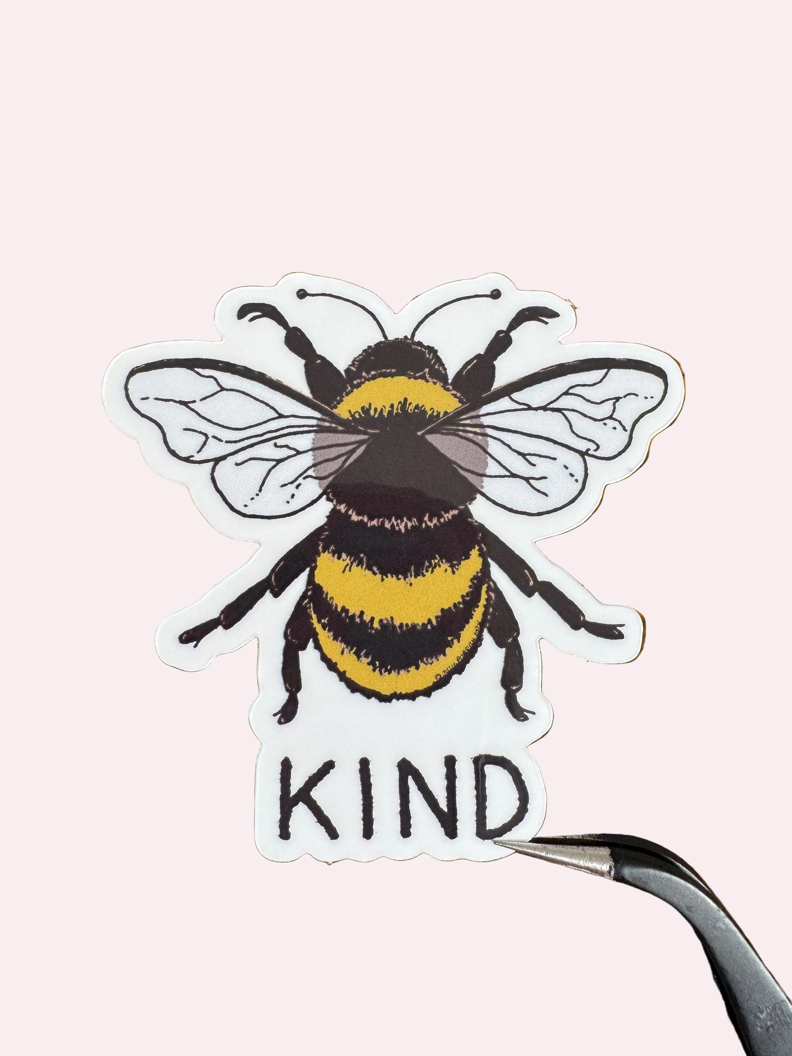 A die-cut sticker featuring a detailed bumblebee and the word 'KIND' in black lettering, held by tweezers on a dark wood background.