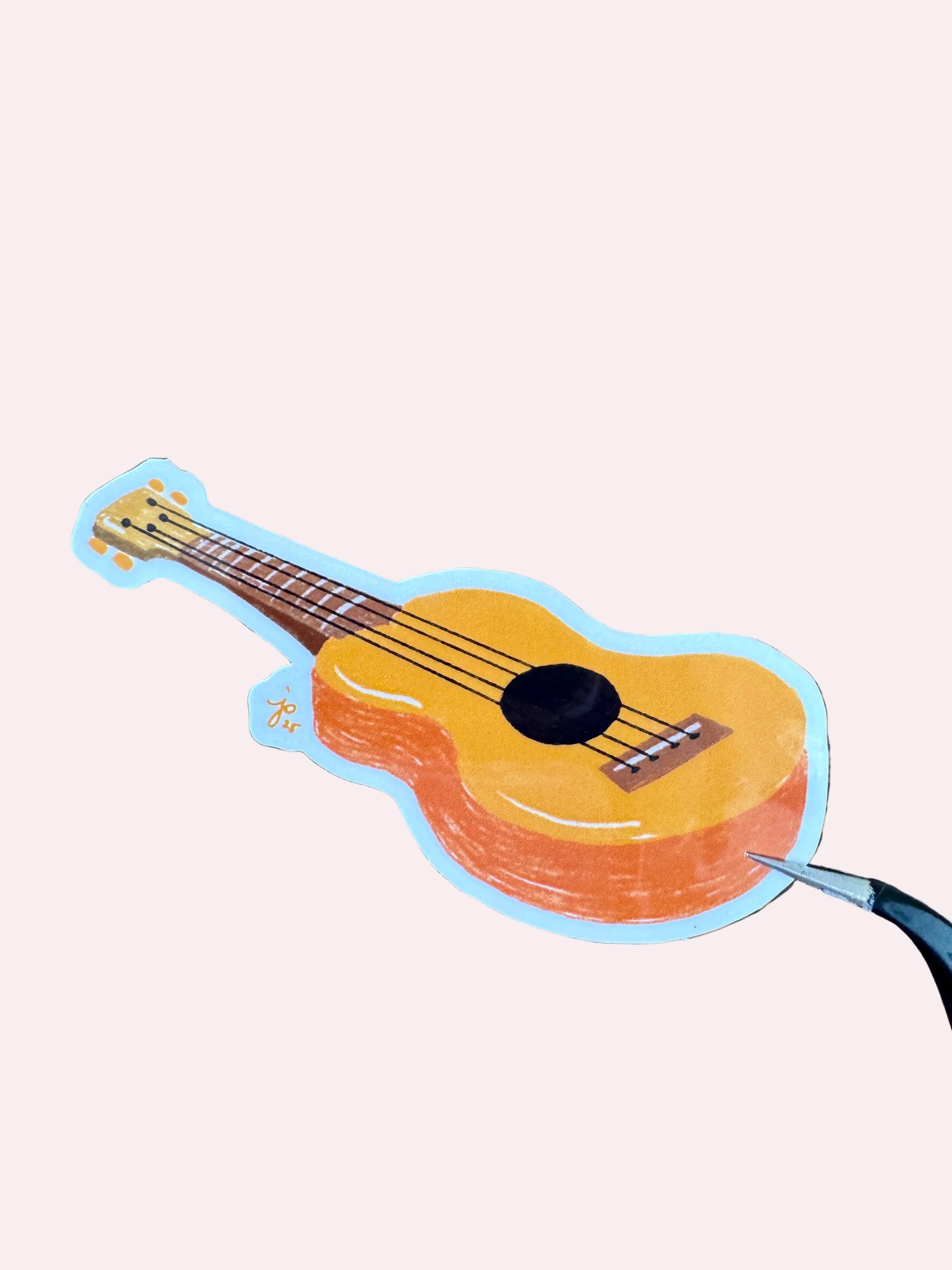 A die-cut sticker of a yellow-orange ukulele with a light blue outline, shown on a dark wood-grain background with a pair of scissors touching the bottom right edge.