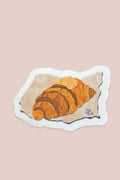 A die-cut sticker featuring an illustration of a croissant on a piece of parchment paper, placed on a dark wood-grained surface.