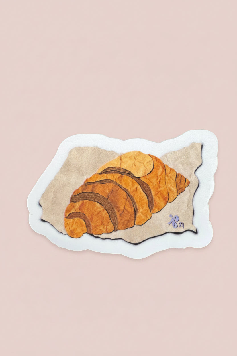 A die-cut sticker featuring an illustration of a croissant on a piece of parchment paper, placed on a dark wood-grained surface.